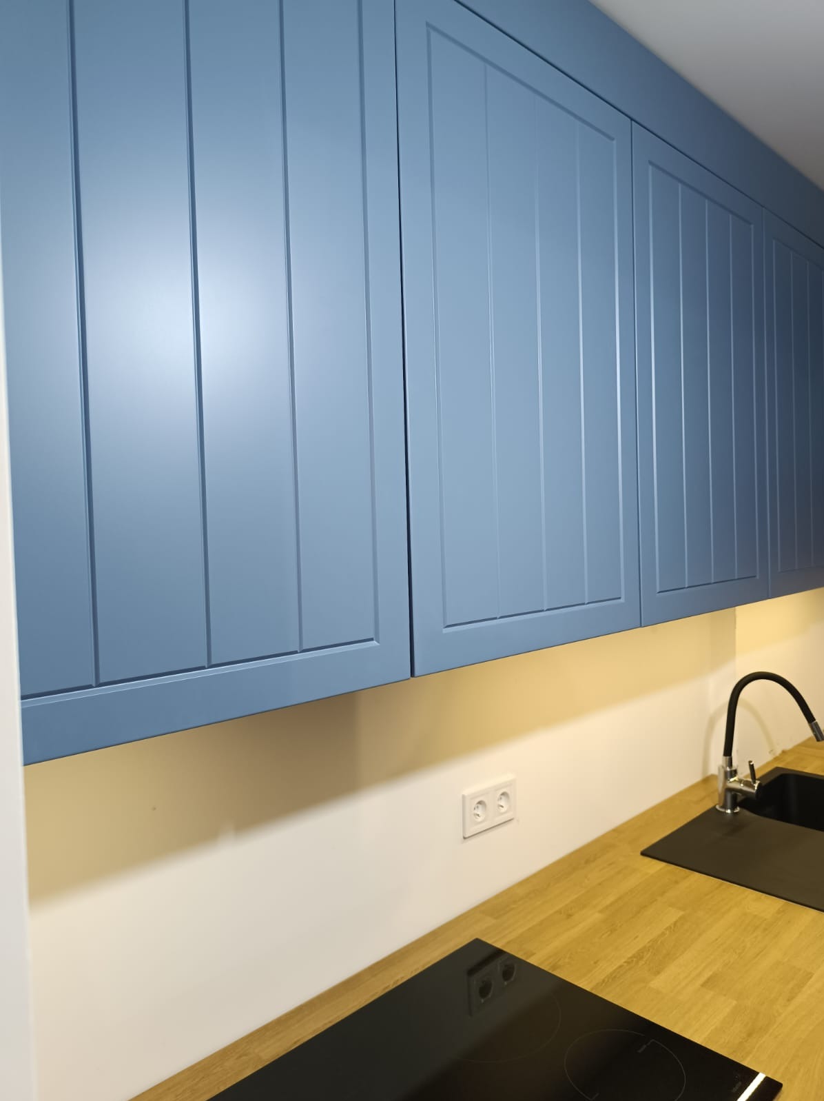 Błękitne, drewniane szafki kuchenne z frezowanymi frontami, oświetlenie podszafkowe, blat z drewna klejonego, czarny zlew i płyta indukcyjna.