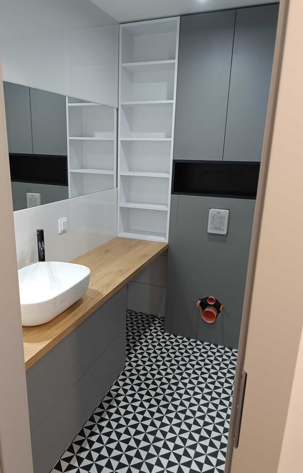 Nowoczesna łazienka z szarą zabudową, drewnianym blatem pod umywalką nablatową i wzorzystą czarno-białą podłogą. Na ścianie widoczny panel sterujący i instalacja kanalizacyjna.