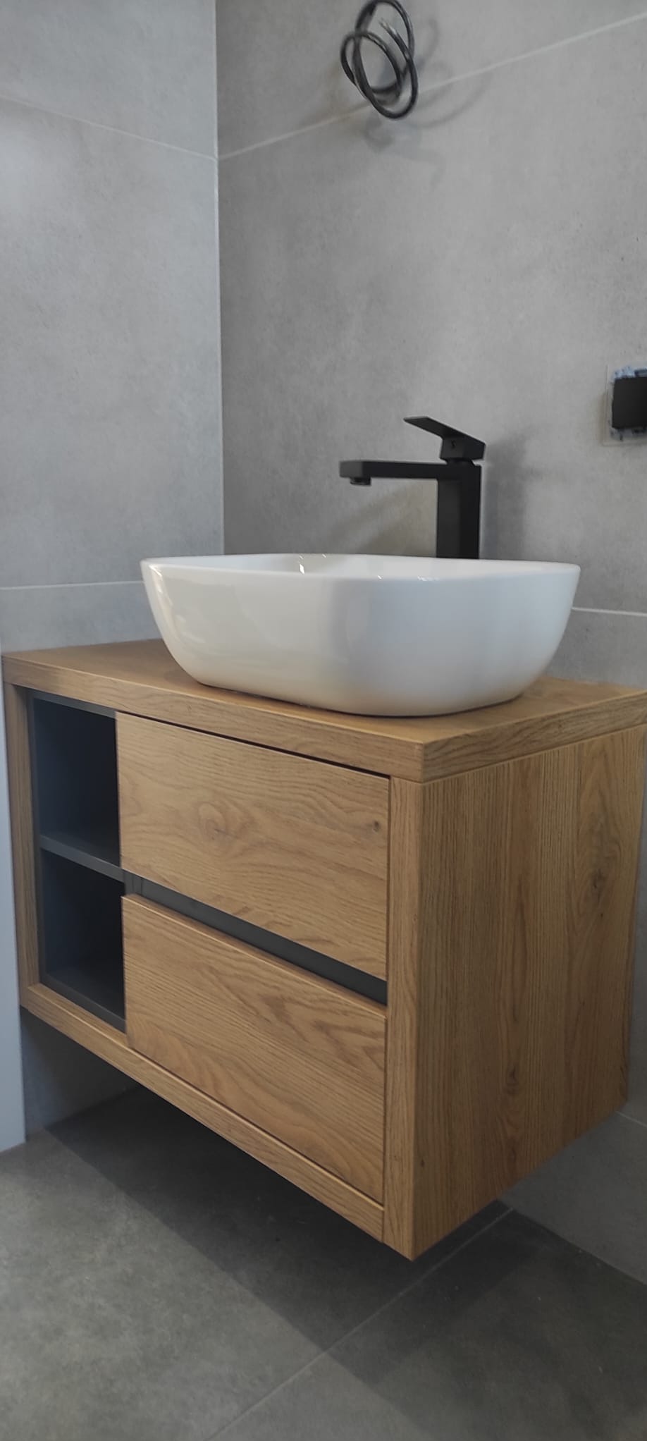 Nowoczesna łazienka z drewnianą szafką pod umywalkę nablatową, czarna bateria i szare płytki na ścianie.