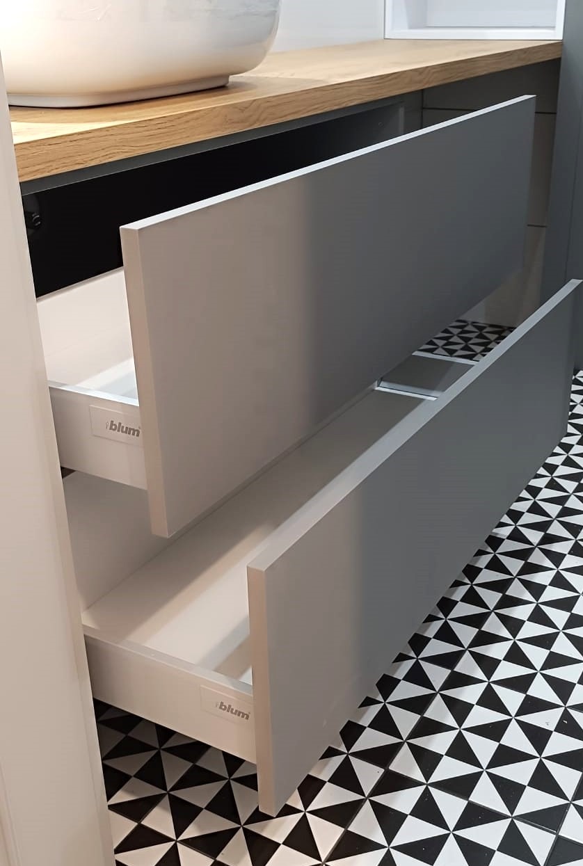 Otwarta szuflada pod umywalką z szarym frontem, widoczny biały korpus i logo Blum, na podłodze czarno-biała mozaika.