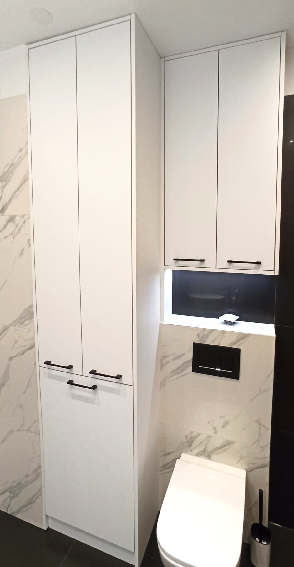 Białe szafki łazienkowe z czarnymi uchwytami na tle marmurowej ściany, widoczna toaleta i czarny panel sterujący spłuczką.