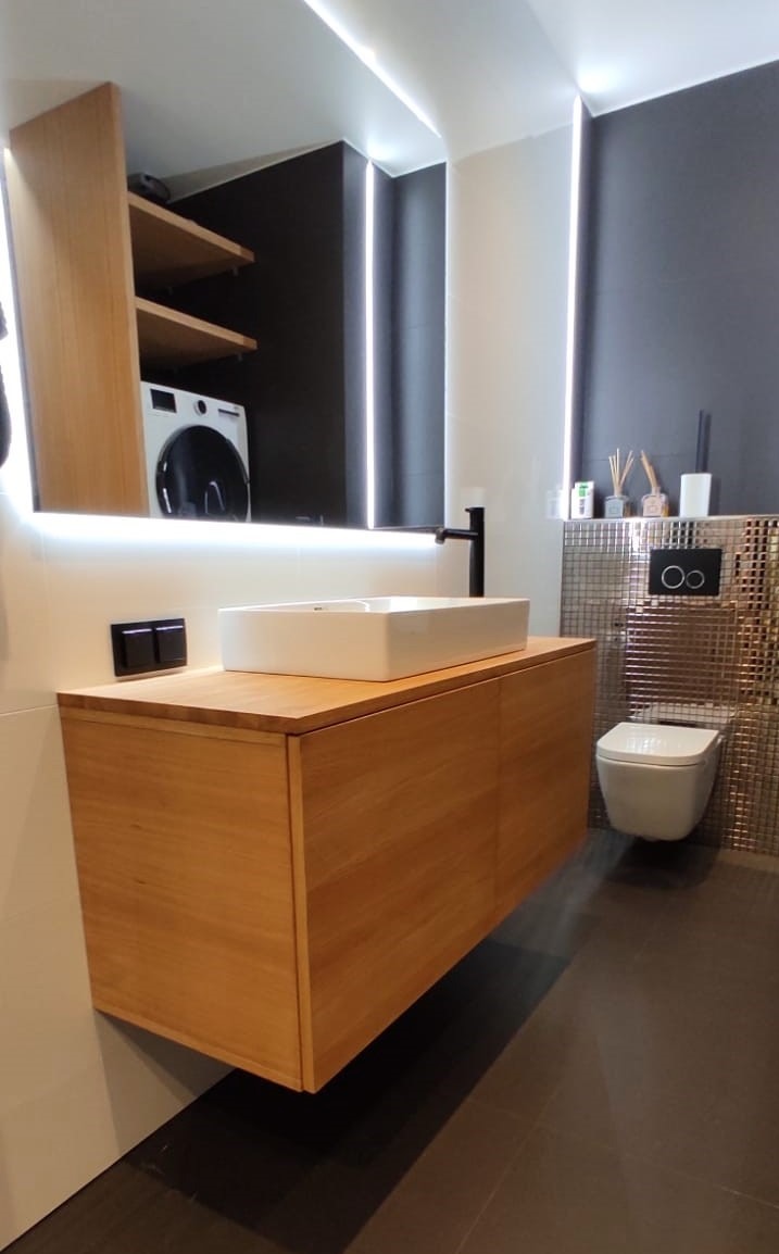 Nowoczesna łazienka z wiszącą szafką z jasnego drewna, białą umywalką nablatową i podświetlanym lustrem. W tle widoczna toaleta i pralka.