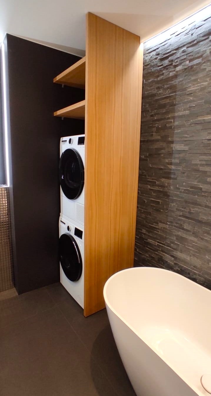 Nowoczesna łazienka z pralką i suszarką w zabudowie z jasnego drewna, obok wolnostojąca wanna i ściana wyłożona kamieniem dekoracyjnym.