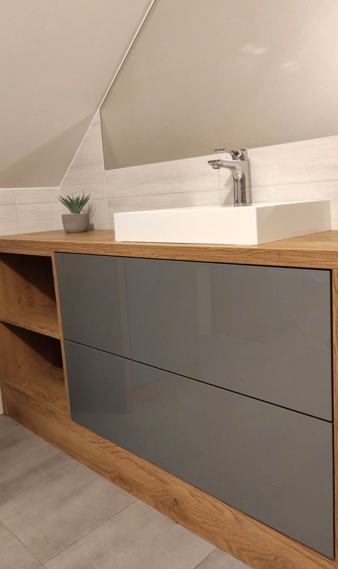 Szara szafka pod umywalkę z drewnianym blatem i otwartymi półkami w łazience na poddaszu, biała prostokątna umywalka nablatowa i minimalistyczna bateria.