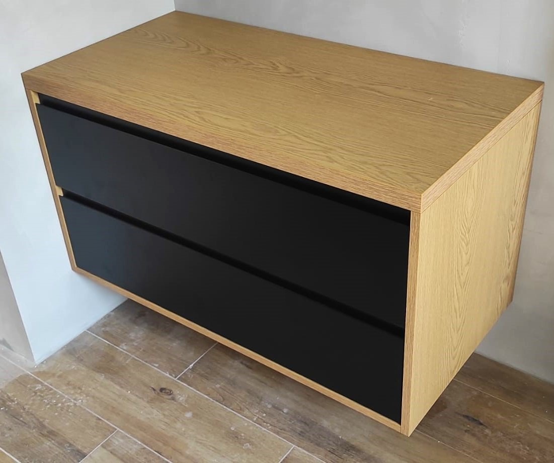 Wisząca szafka z dwoma czarnymi szufladami i korpusem z jasnego drewna, widoczna na tle jasnej ściany i podłogi z płytek imitujących drewno.