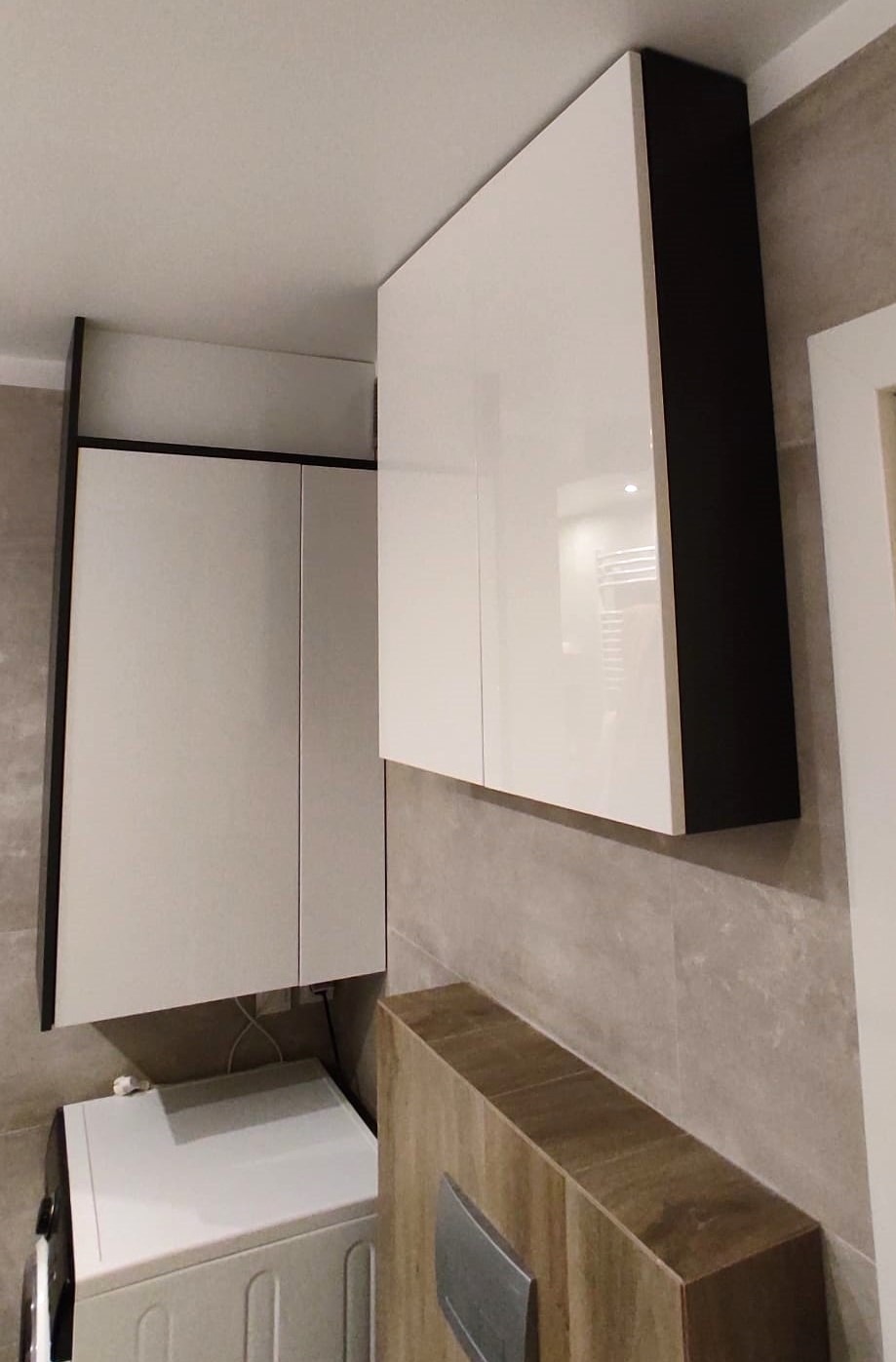 Białe, lakierowane szafki wiszące z czarnymi obrzeżami w łazience, zamontowane nad pralką i zabudową umywalkową z szarej płytki imitującej kamień.