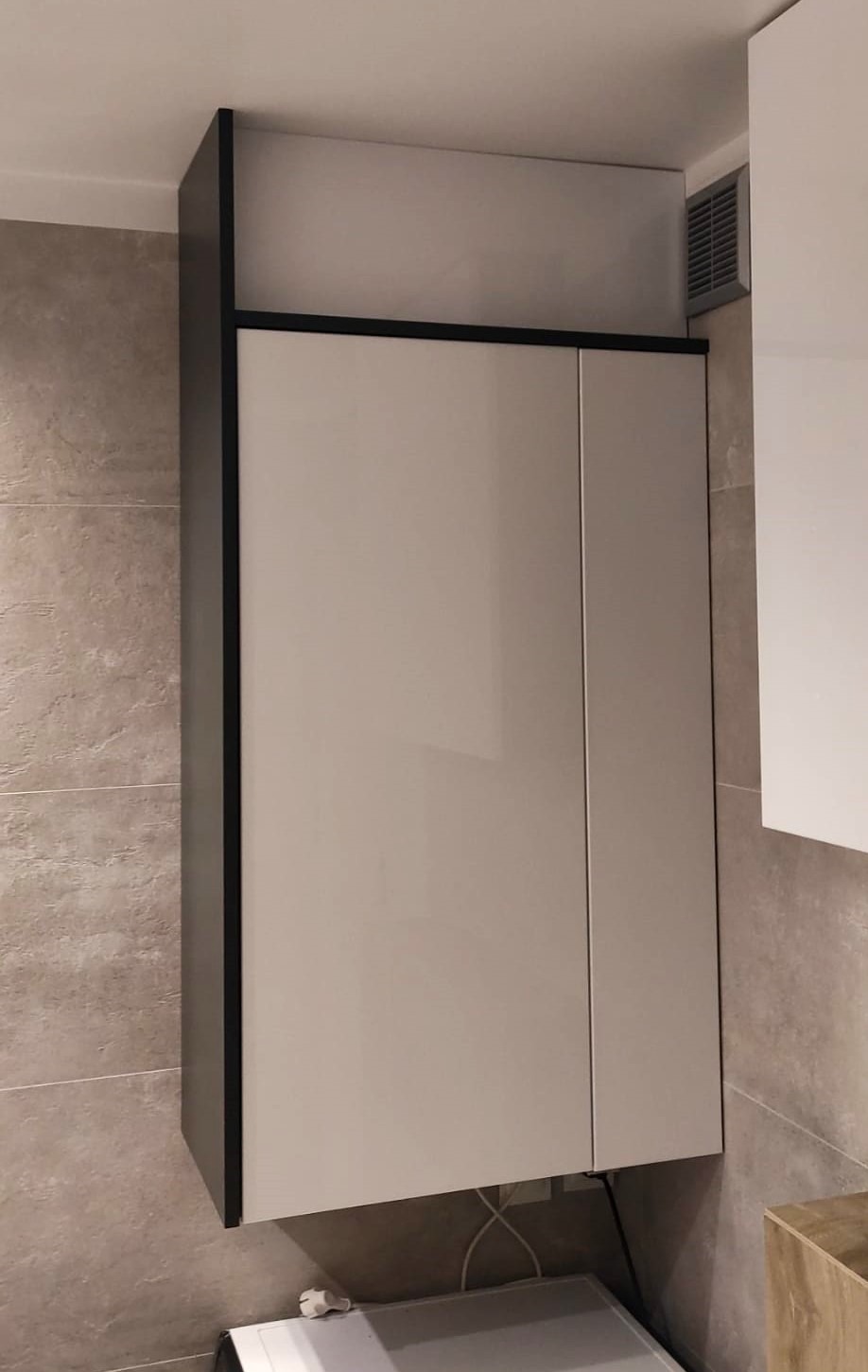 Szara szafka kuchenna wisząca, z czarnymi obramowaniami, zamontowana na ścianie z płytek imitujących kamień, widoczna kratka wentylacyjna w suficie.