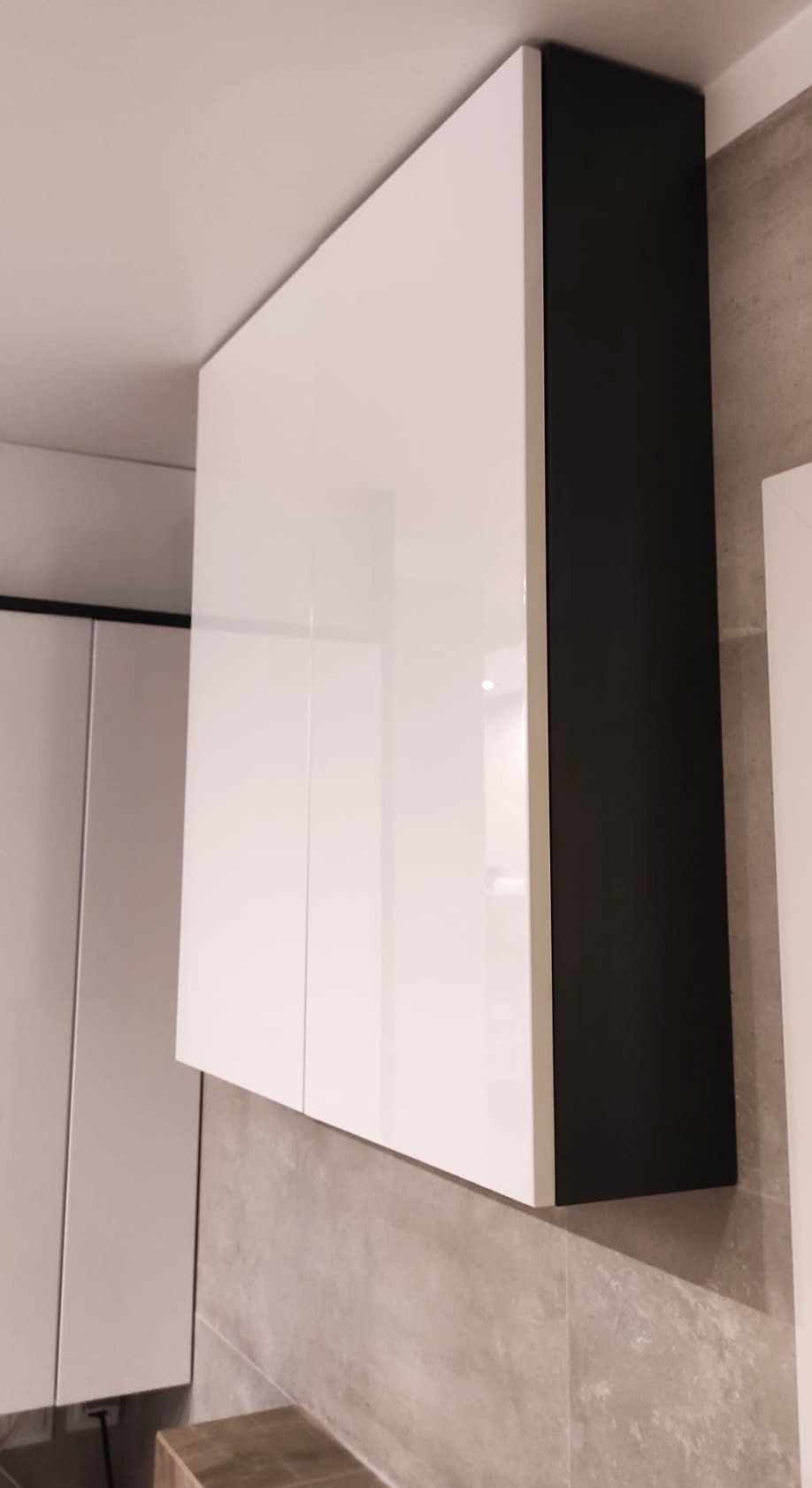 Biała szafka wisząca z czarnym bokiem, zamontowana na ścianie wyłożonej szarymi płytkami, widoczna z perspektywy dolnej.