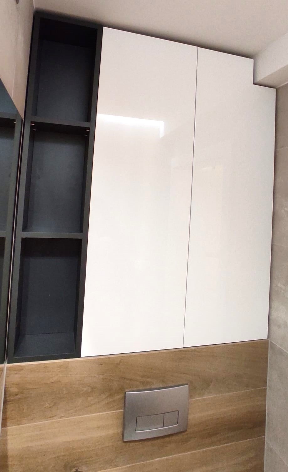 Nowoczesna szafka łazienkowa z białymi, lakierowanymi frontami i otwartymi półkami w kolorze grafitowym, zamontowana nad zabudową z drewnopodobnych paneli z widocznym przyciskiem spłuczki.