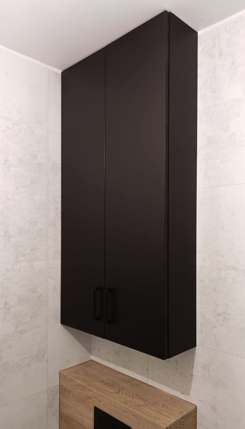 Czarna, wisząca szafka łazienkowa z matowymi frontami i minimalistycznymi uchwytami, zamontowana nad drewnianą szafką w łazience w Gruszowie, na tle ściany wyłożonej płytkami imitującymi marmur.