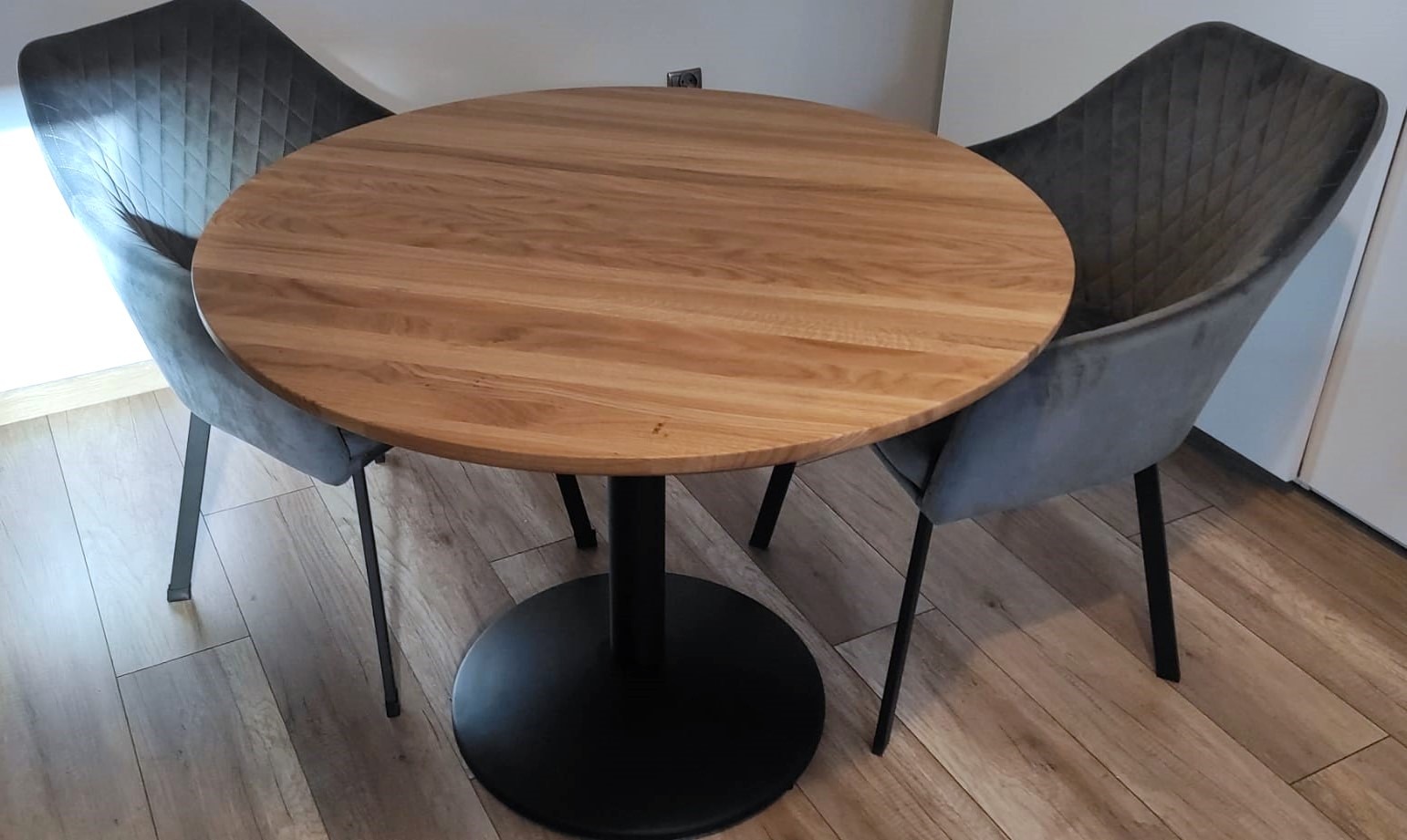 Okrągły stół z blatem z jasnego drewna na czarnej, metalowej nodze, otoczony dwoma szarymi, tapicerowanymi krzesłami.
