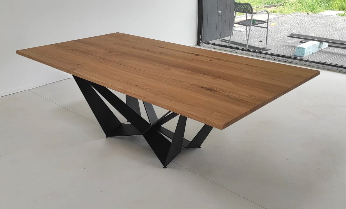 Stół z blatem z jasnego drewna i geometryczną podstawą z czarnych, metalowych elementów, ustawiony na białej podłodze, z widokiem na taras i krzesło przez przeszklone drzwi.