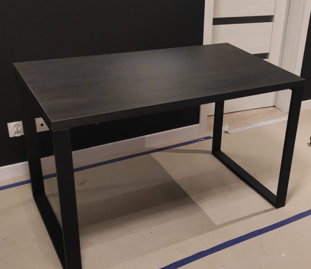 Stół o czarnym, metalowym stelażu i blacie w odcieniach szarości, ustawiony na podłodze zabezpieczonej taśmą malarską, na tle czarnej ściany i białych drzwi.