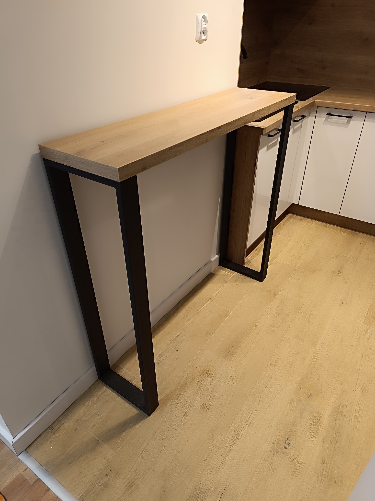 Blat z jasnego drewna na czarnych, metalowych nogach, przylegający do ściany w kuchni z białymi szafkami.