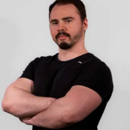 Mężczyzna z brodą, w czarnej koszulce, z założonymi rękami na tle jasnej ściany.