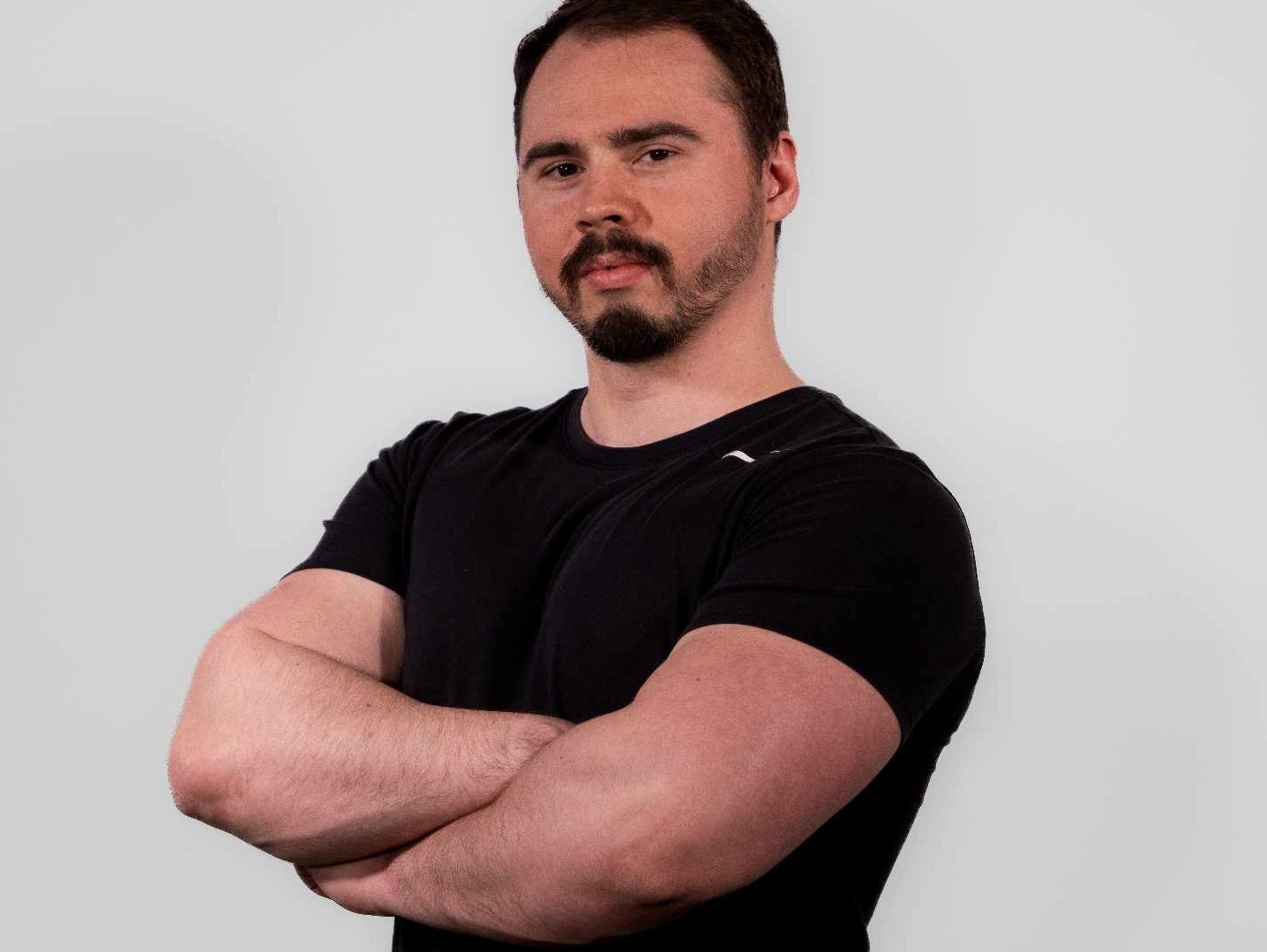Mężczyzna z brodą, w czarnej koszulce, z założonymi rękami na tle jasnej ściany.