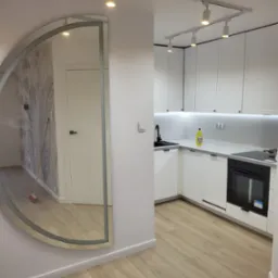 Nowoczesna, jasna kuchnia z białymi szafkami, drewnianą podłogą i dużym, półokrągłym lustrem na ścianie, odbijającym fragment pokoju z tapetą w motywy roślinne i stojącą miotłą.