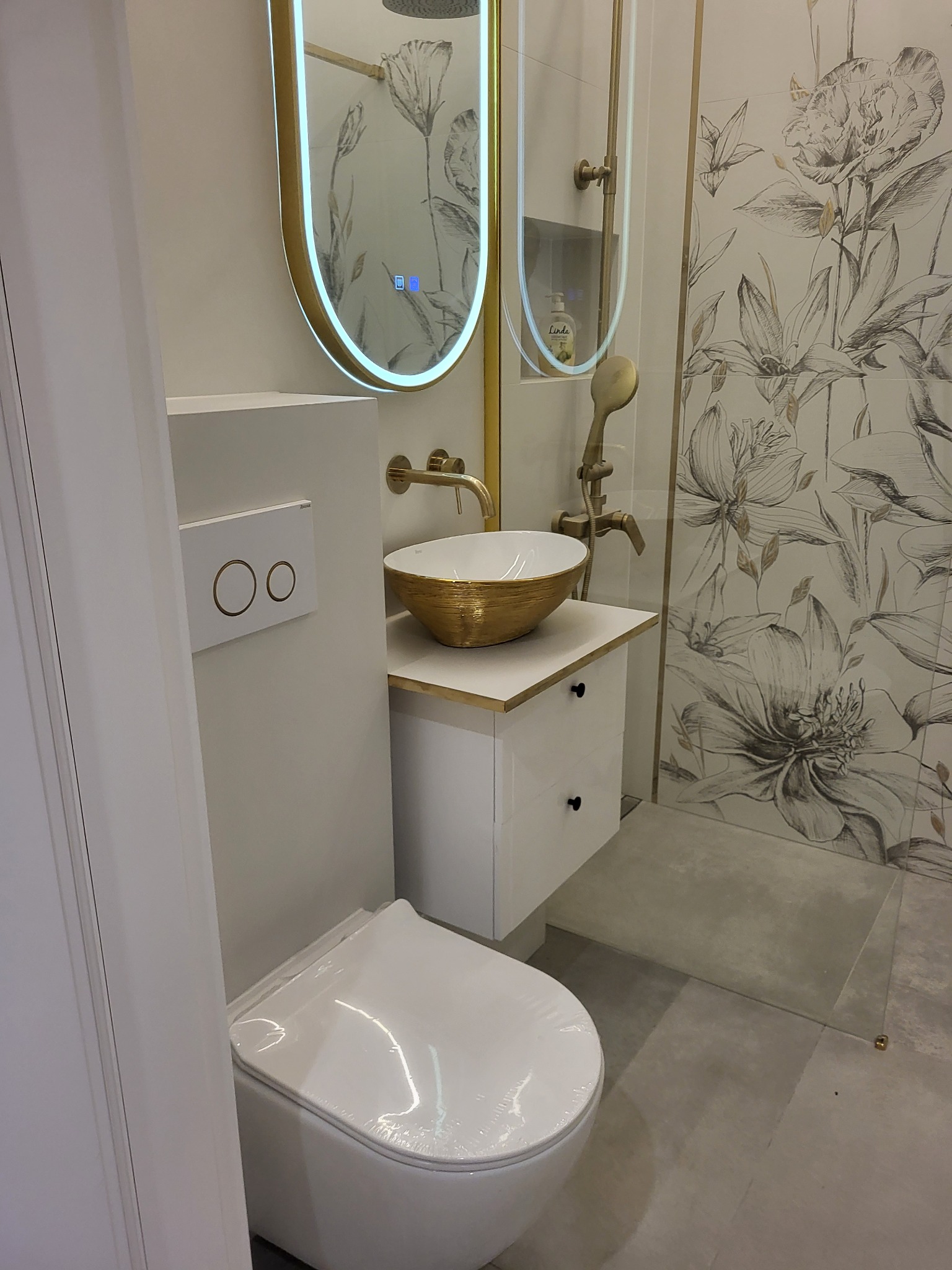 Nowoczesna łazienka z umywalką w kolorze złota, armaturą w odcieniach złota, toaletą z białym panelem spłukującym i kabiną prysznicową z motywem kwiatowym na ścianie.