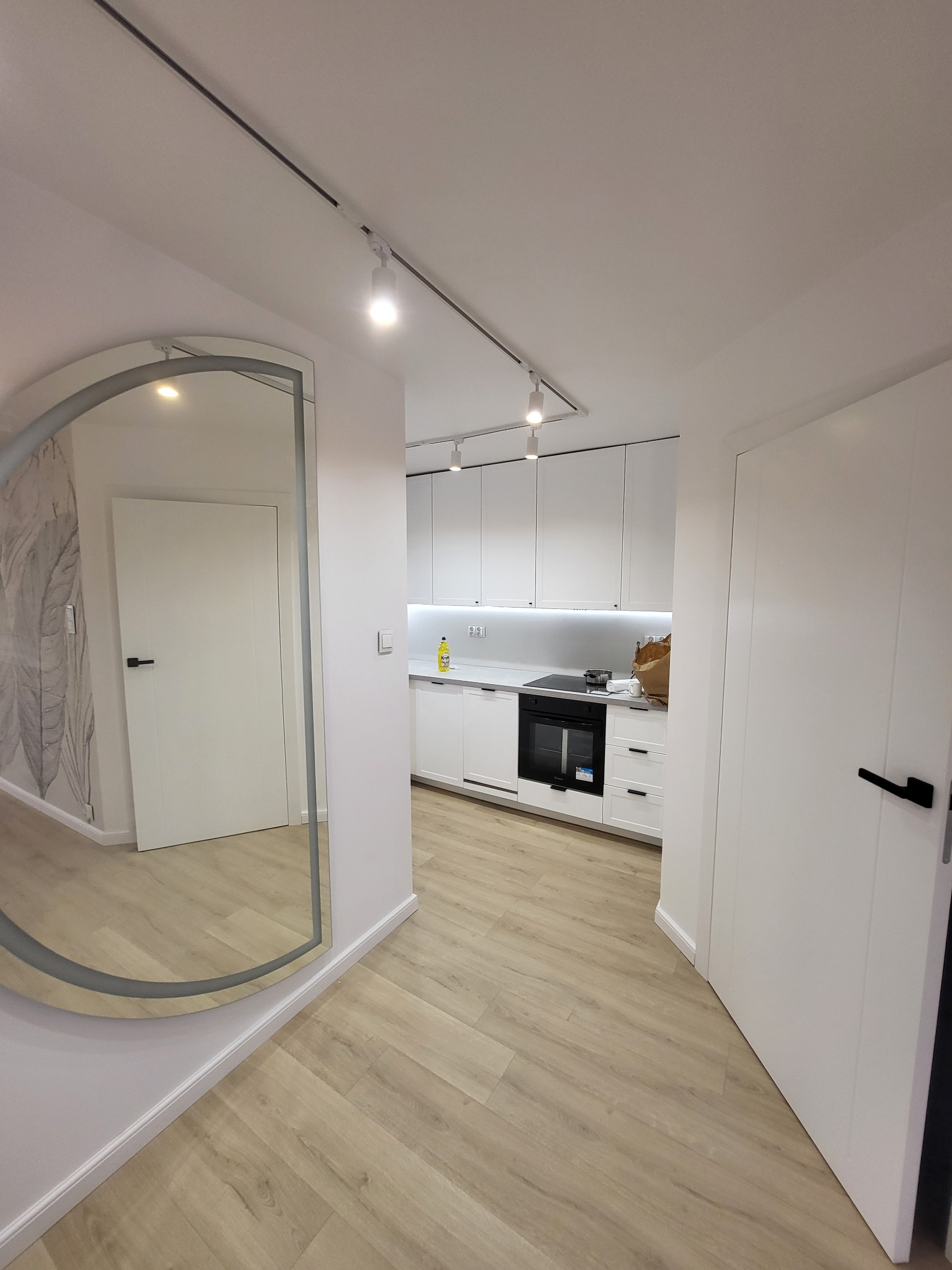 Nowoczesne wnętrze mieszkania z białymi meblami kuchennymi, drewnopodobną podłogą ułożoną w jodełkę i dużym lustrem w przedpokoju, oświetlone reflektorami.