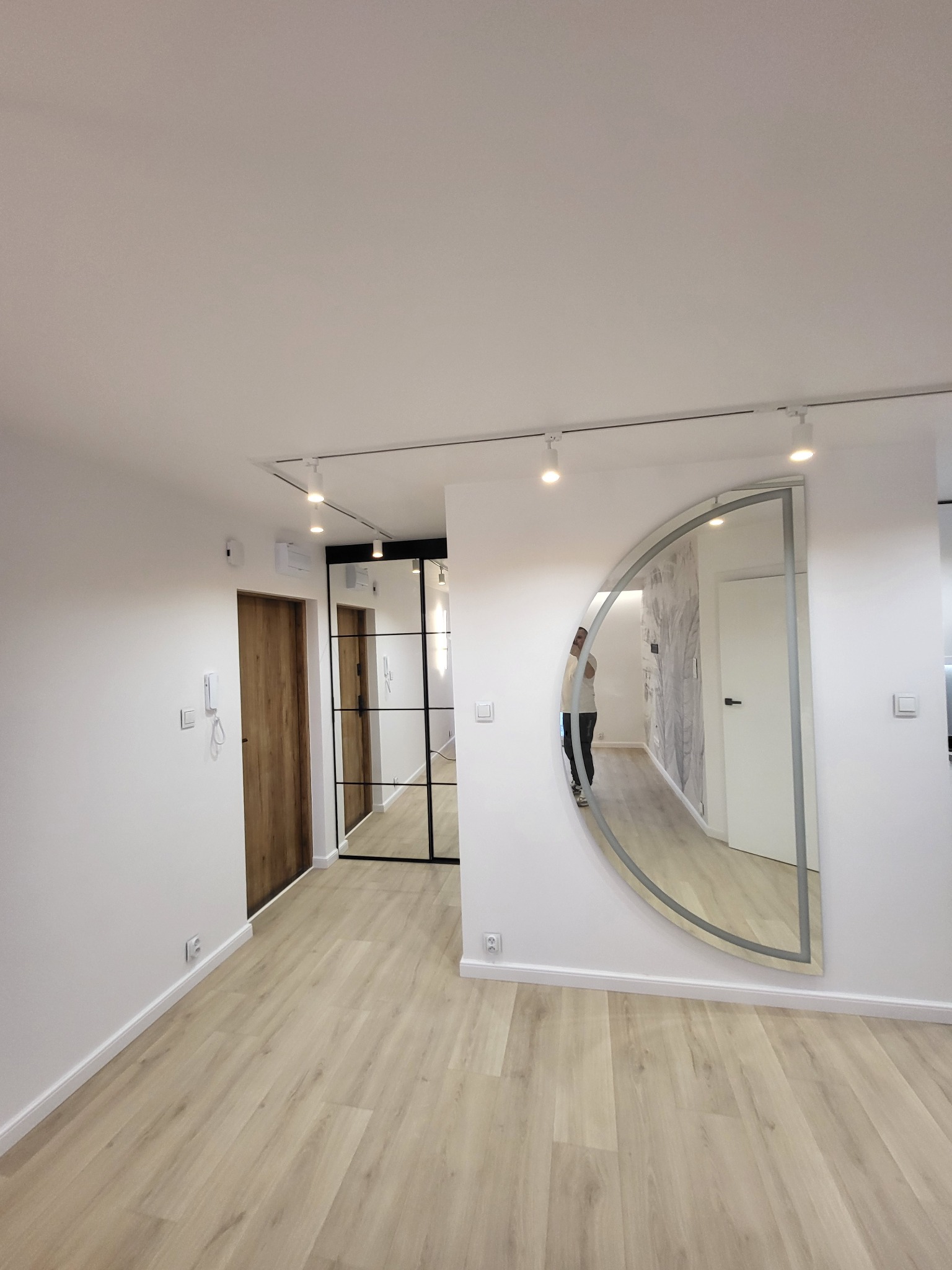 Jasne wnętrze mieszkania z panelami podłogowymi, białymi ścianami, drewnianymi drzwiami, przeszklonymi drzwiami w czarnej ramie i półokrągłym lustrem z widocznym odbiciem osoby.