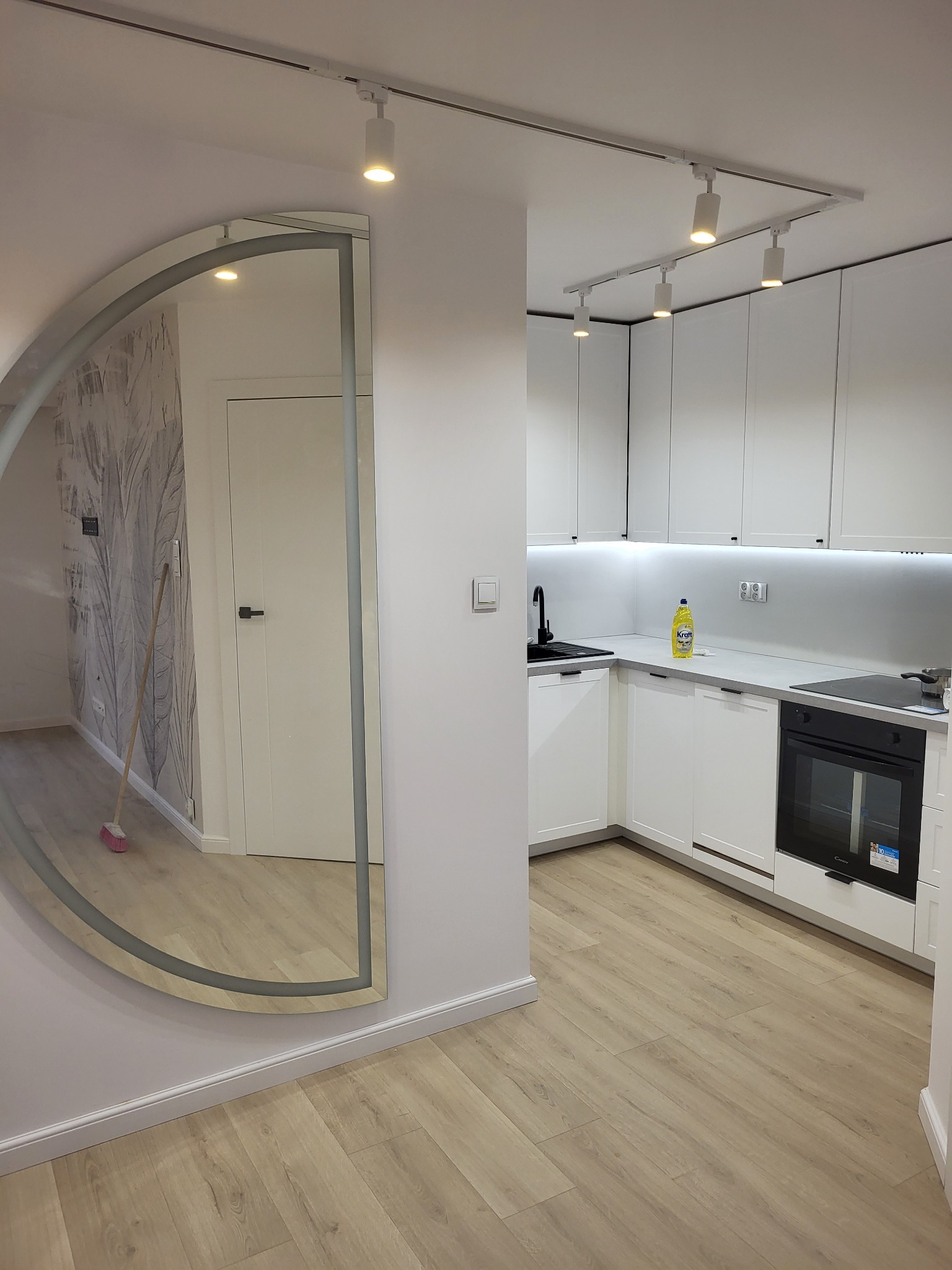 Nowoczesna, jasna kuchnia z białymi szafkami, drewnianą podłogą i dużym, półokrągłym lustrem na ścianie, odbijającym fragment pokoju z tapetą w motywy roślinne i stojącą miotłą.