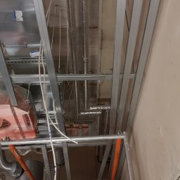A&W SERVICE Wiesław Iwanowski - Montaż instalacji wentylacyjnej: widok na sufit z widocznymi przewodami wentylacyjnymi w izolacji, metalową konstrukcją podwieszaną, rurami z tworzywa i elementami rusztowania.