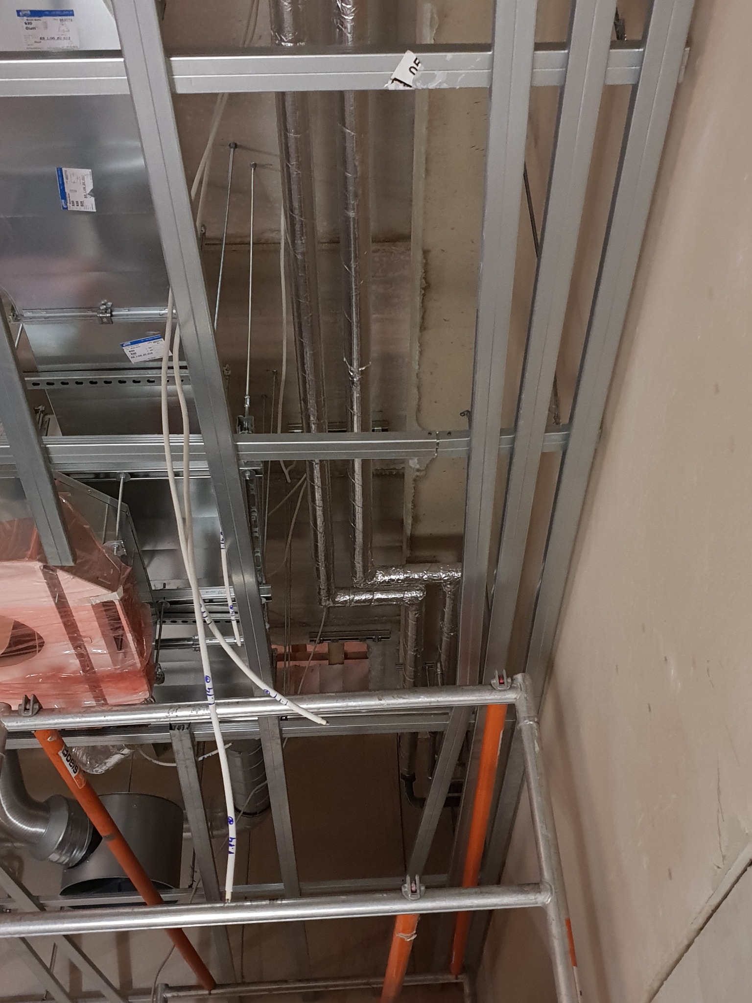 Montaż instalacji wentylacyjnej: widok na sufit z widocznymi przewodami wentylacyjnymi w izolacji, metalową konstrukcją podwieszaną, rurami z tworzywa i elementami rusztowania.