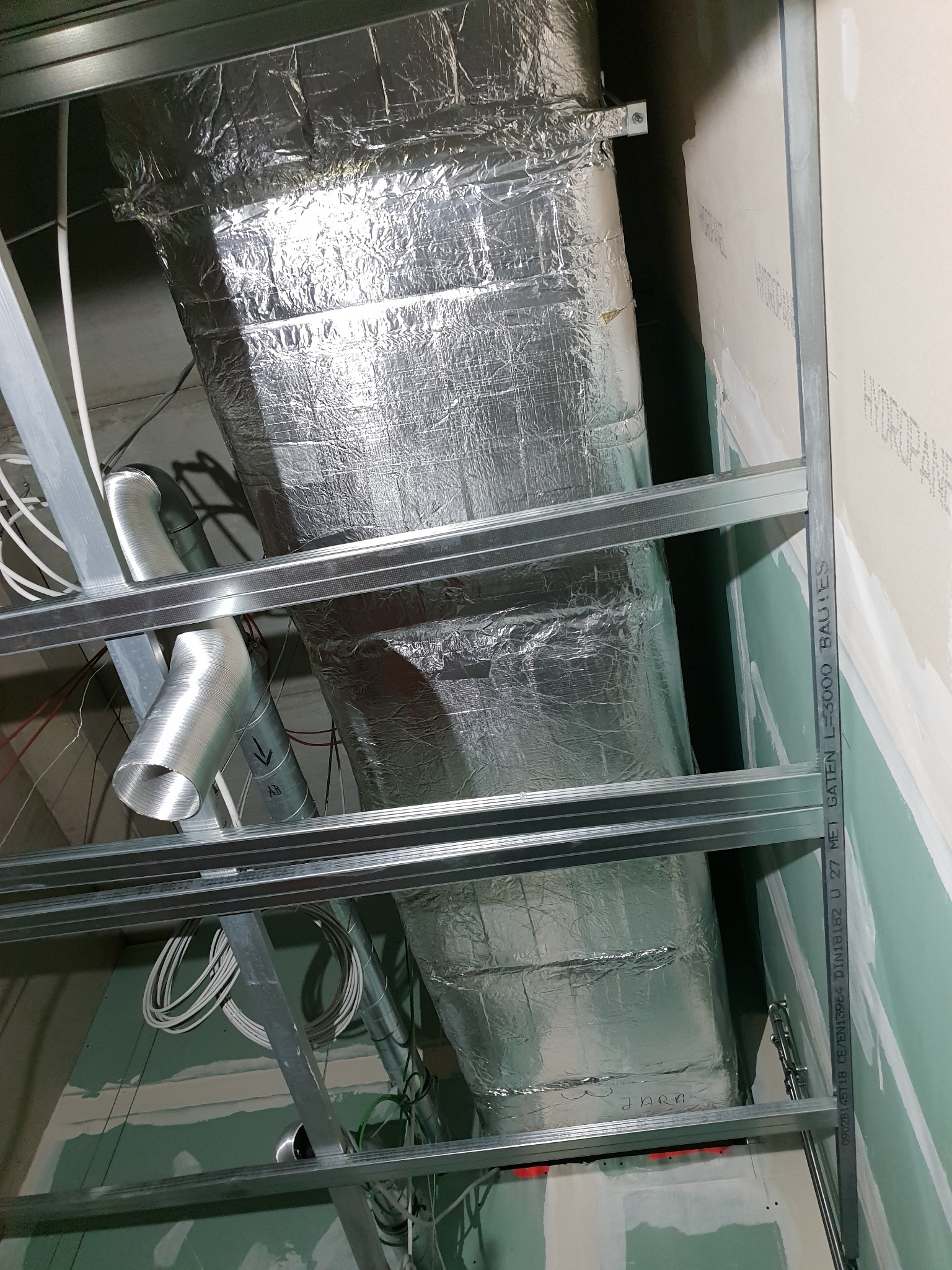 Kanciasty kanał wentylacyjny owinięty folią aluminiową, zamontowany pod sufitem z metalową konstrukcją i płytami gipsowo-kartonowymi, widoczne przewody i elastyczny przewód wentylacyjny.