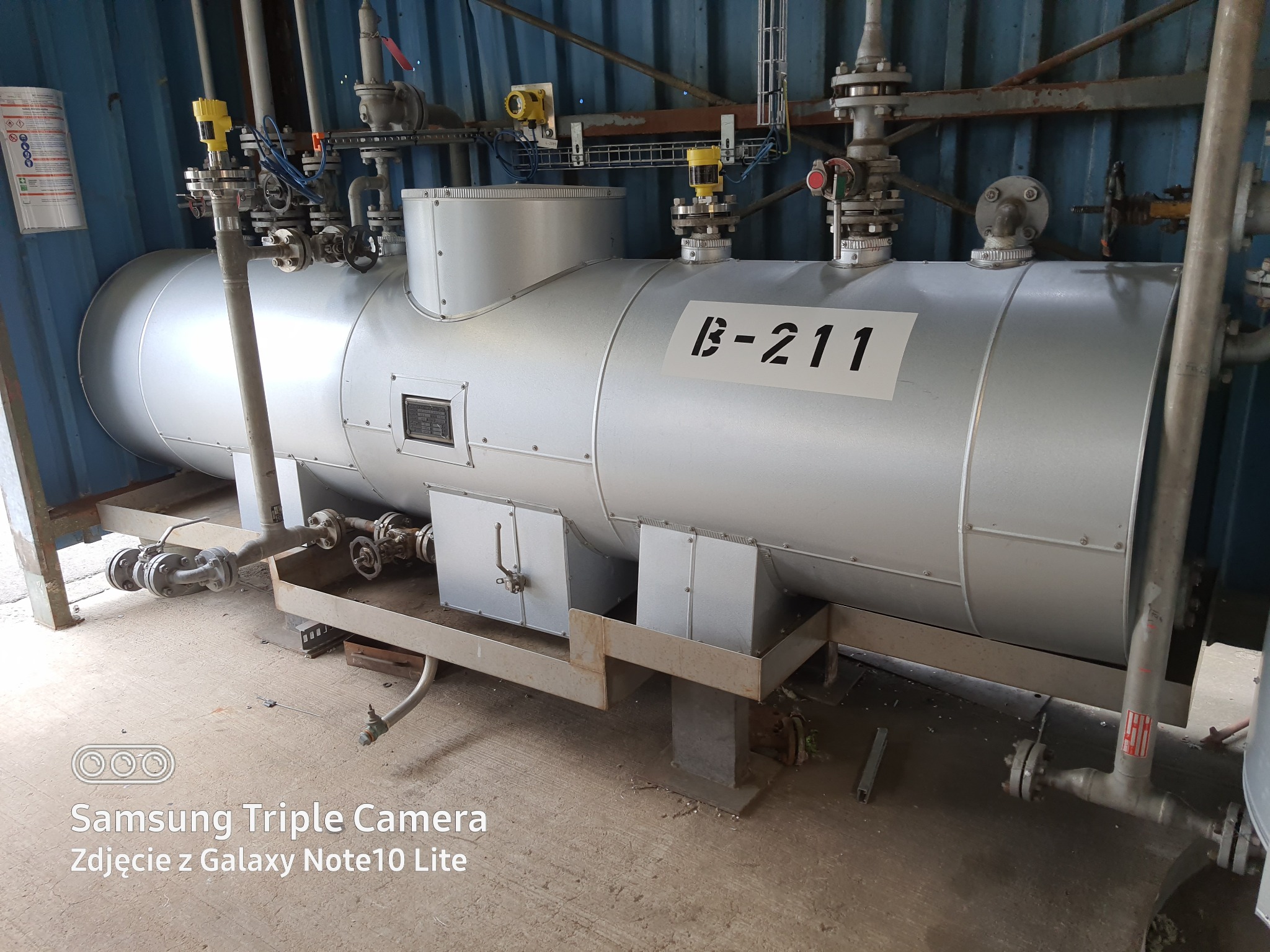 Srebrny, poziomy zbiornik B-211 z licznymi rurami, zaworami i czujnikami w przemysłowym otoczeniu z blachy falistej.