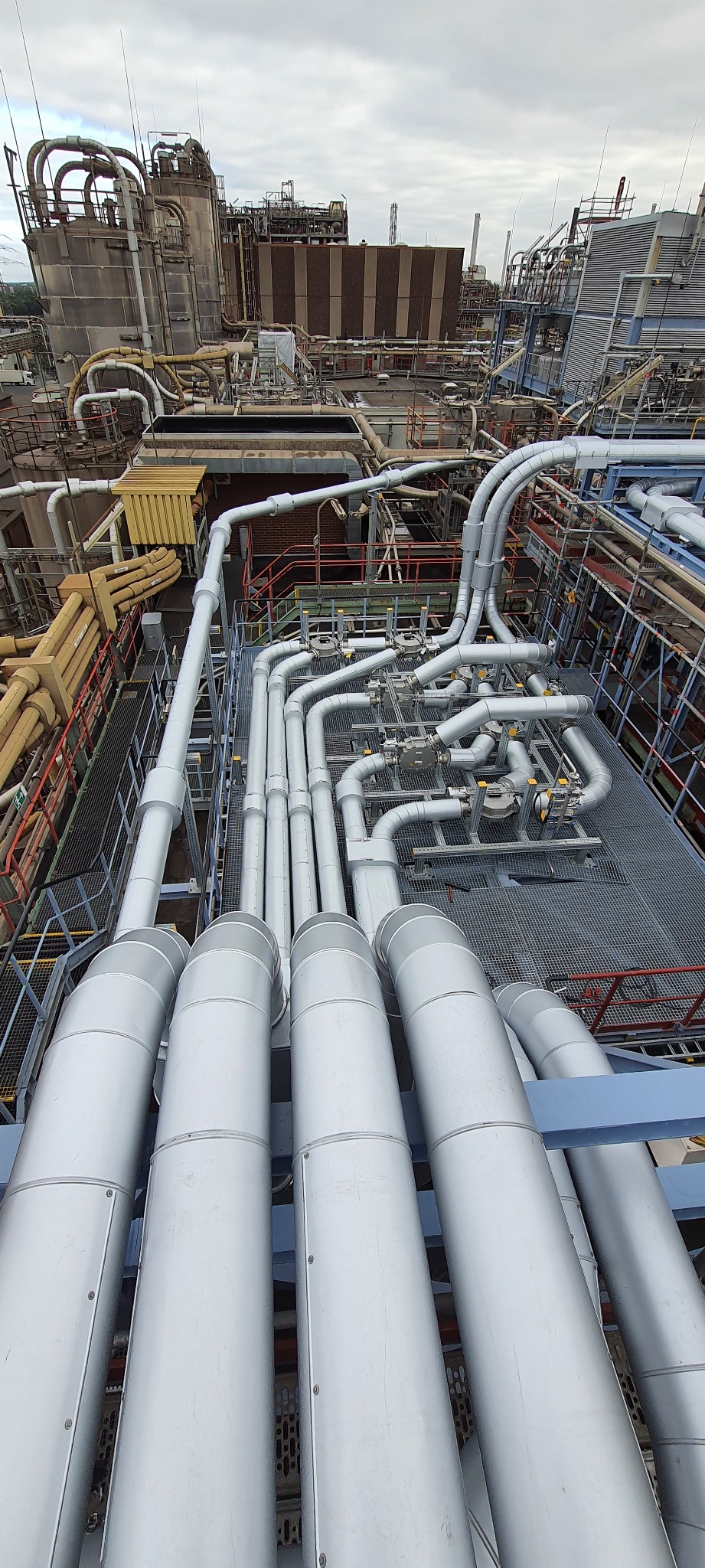 Widok z góry na skomplikowany system rur przemysłowych w fabryce, z licznymi złączkami, zaworami i izolacją, na tle budynków fabrycznych i pochmurnego nieba.