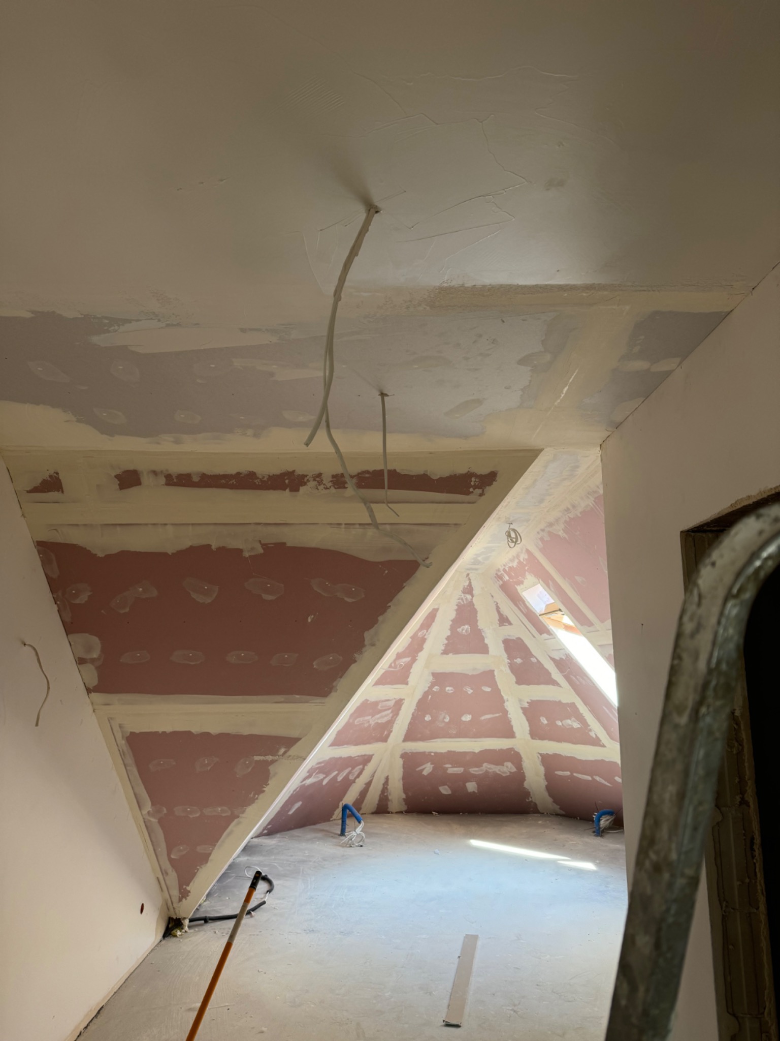 Poddasze w trakcie prac wykończeniowych, widoczne płyty gipsowo-kartonowe na skosach ścian, częściowo pokryte masą szpachlową, wystające kable, drabina w kadrze.