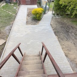 OLEH-BUD OLEH ULEI - Widok z góry na nowo wyłożony chodnik z szarej kostki brukowej, prowadzący od drewnianych schodów do furtki w ogrodzeniu. Po lewej stronie trawnik z pojedynczymi płytami chodnikowymi, po prawej...