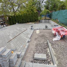 OLEH-BUD OLEH ULEI - Układanie szarej kostki brukowej na podwórku, widoczna ubijarka w folii, stosy kostki, częściowo ułożona nawierzchnia i przygotowane podłoże z kamieni i piasku.