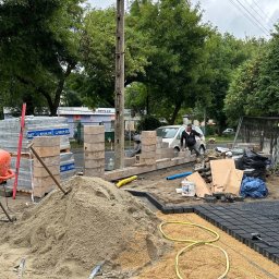 OLEH-BUD OLEH ULEI - Budowa murku z betonowych bloczków przy chodniku, widoczna betoniarka, palety z bloczkami Bruk-Bet, piasek, kostka brukowa i pracownicy w trakcie układania.