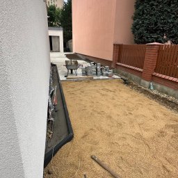 OLEH-BUD OLEH ULEI - Przygotowanie terenu pod kostkę brukową: wyrównany piasek, obrzeża, paleta z kostką, taczka i młotek brukarski na budowie między budynkami.