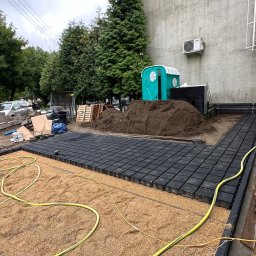 OLEH-BUD OLEH ULEI - Układanie czarnych, kwadratowych płyt na piasku i żwirze przed budynkiem, widoczna sterta ziemi i przenośna toaleta w tle.