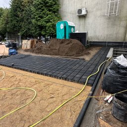 OLEH-BUD OLEH ULEI - Układanie czarnej kostki brukowej na podsypce z piasku na zewnątrz budynku, widoczny przenośny WC i przygotowany teren pod dalsze prace.