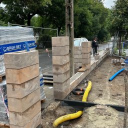 OLEH-BUD OLEH ULEI - Budowa niskiego murku z betonowych bloczków na zewnątrz, widoczne rury karbowane w kolorze niebieskim i żółtym, paleta z kostką brukową w tle.