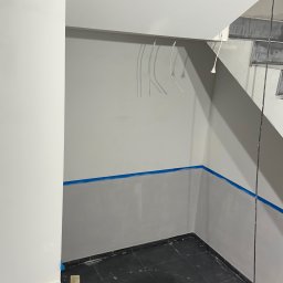 OLEH-BUD OLEH ULEI - Przygotowanie do malowania: ściany zabezpieczone taśmą malarską, widoczne przewody elektryczne, czarna podłoga z płytek, narzędzia malarskie obok ściany, pracownik w kombinezonie.