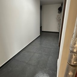 OLEH-BUD OLEH ULEI - Korytarz z szarymi płytkami podłogowymi, świeżo pomalowane białe ściany, widoczne kable i rury w tle, sufit z wbudowanym oświetleniem.