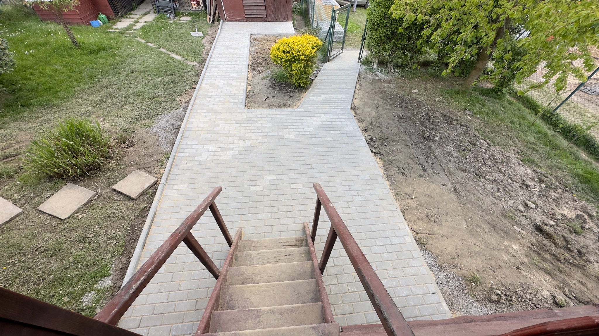 Widok z góry na nowo wyłożony chodnik z szarej kostki brukowej, prowadzący od drewnianych schodów do furtki w ogrodzeniu. Po lewej stronie trawnik z pojedynczymi płytami chodnikowymi, po prawej...