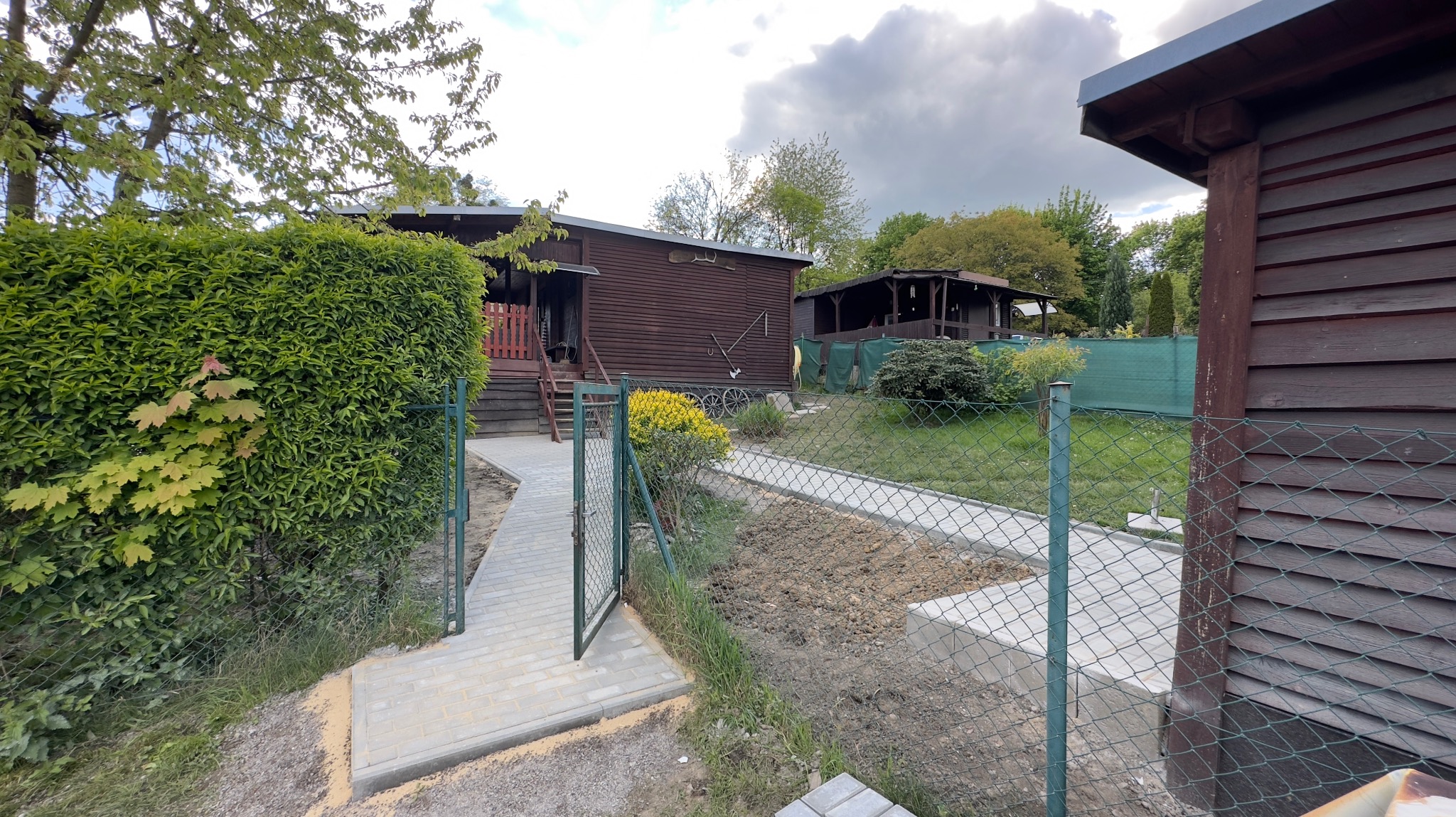 Dwa drewniane domki letniskowe widoczne zza otwartej furtki i ścieżki z kostki brukowej. Jeden domek z werandą i schodami, drugi z zadaszonym tarasem. W tle zielona siatka ogrodzeniowa i drzewa.