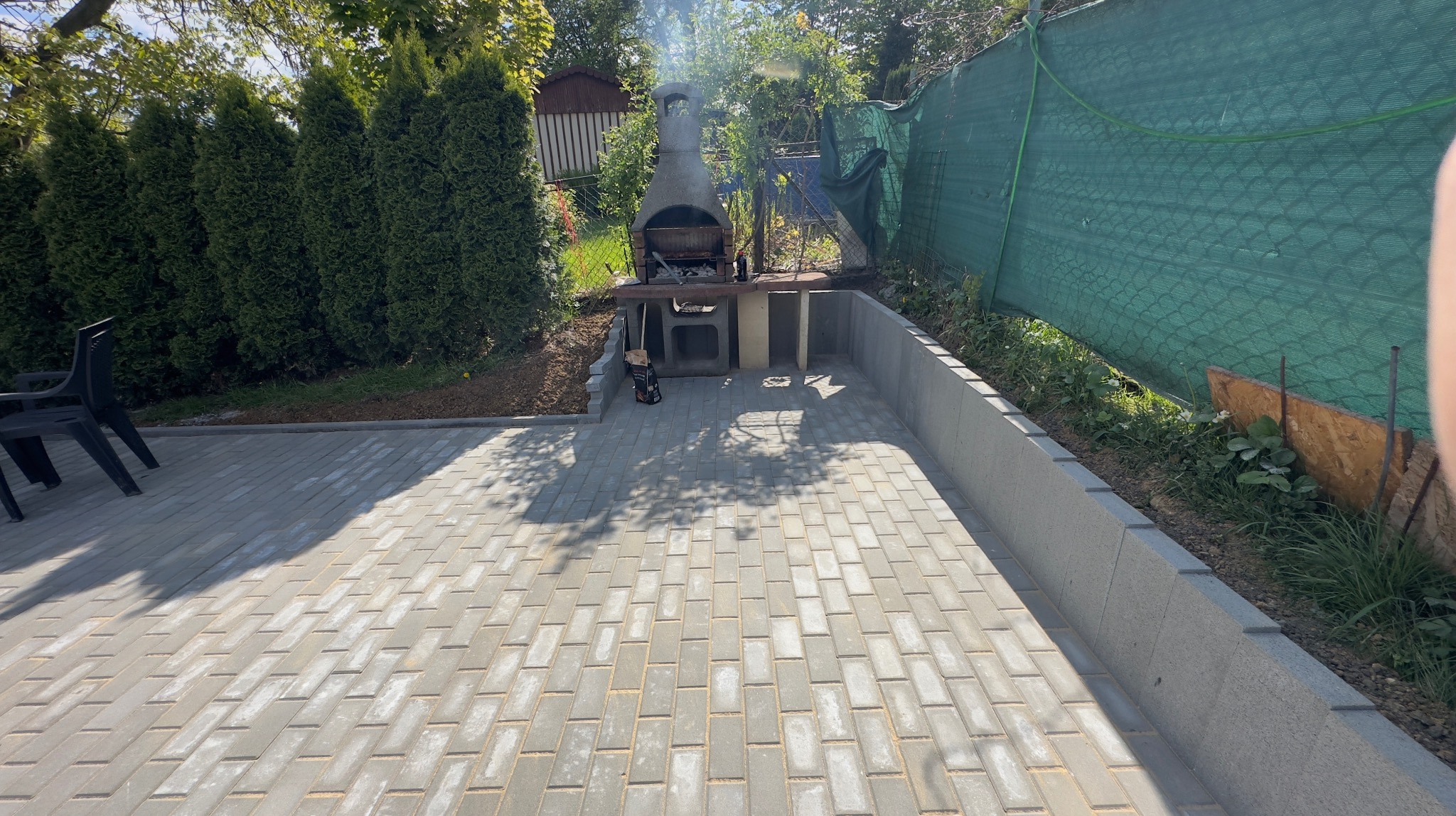 Ułożona szara kostka brukowa na posesji z betonowym grillem ogrodowym z kominem, w tle tuje i siatka maskująca.