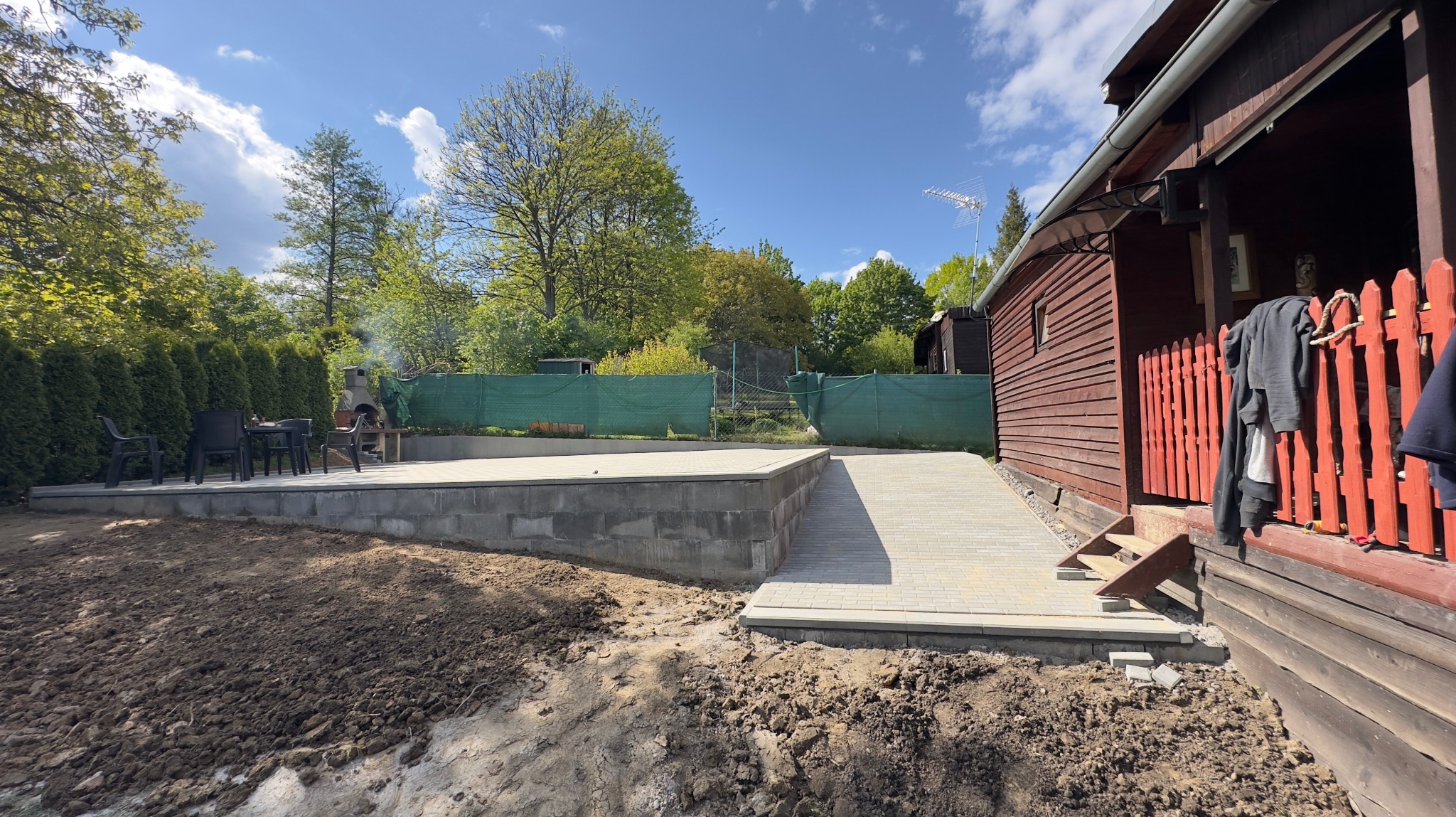 Ułożona kostka brukowa tworząca ścieżkę i taras przy drewnianym domu z czerwonym ogrodzeniem, widoczne przygotowane podłoże i meble ogrodowe w tle, w słoneczny dzień.