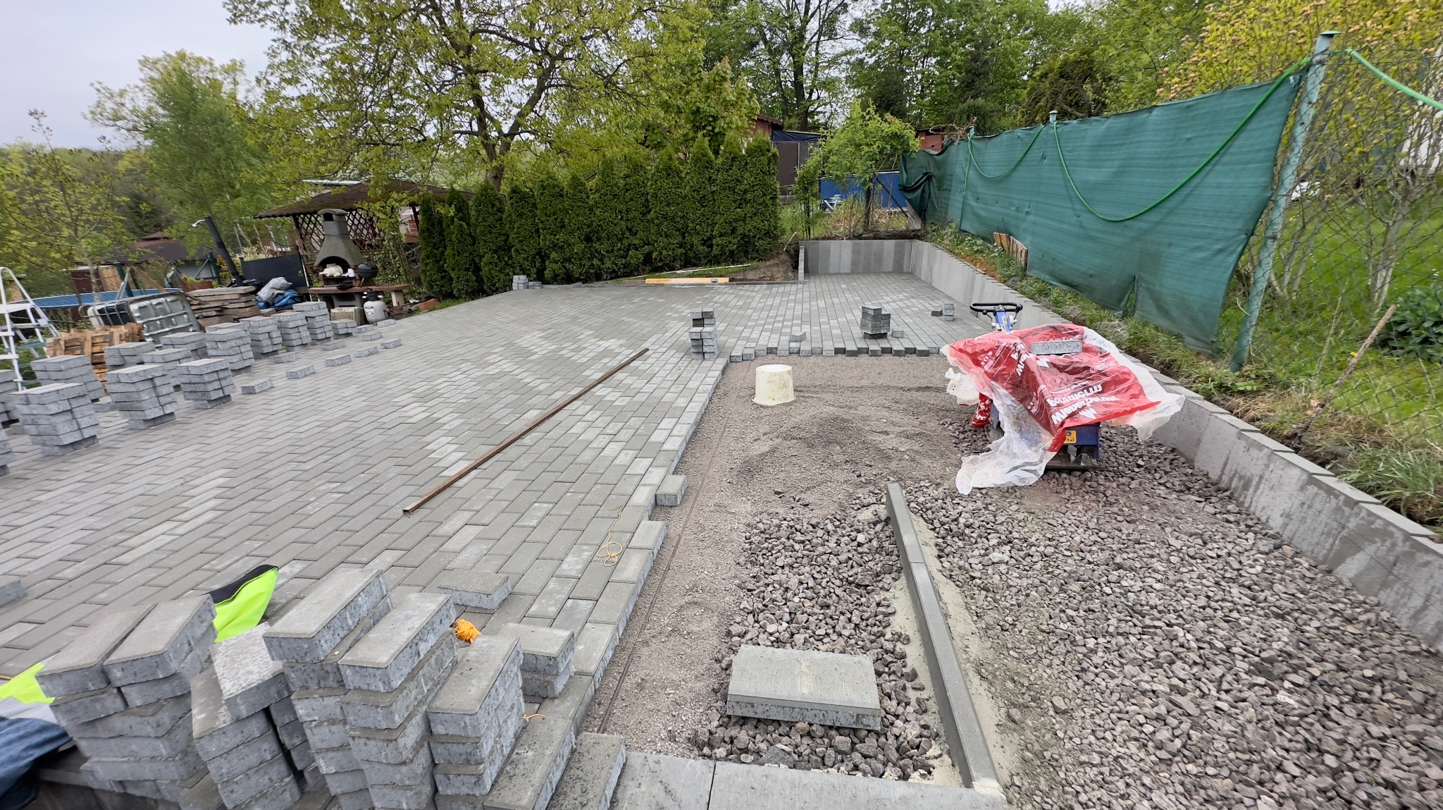 Układanie szarej kostki brukowej na podwórku, widoczna ubijarka w folii, stosy kostki, częściowo ułożona nawierzchnia i przygotowane podłoże z kamieni i piasku.