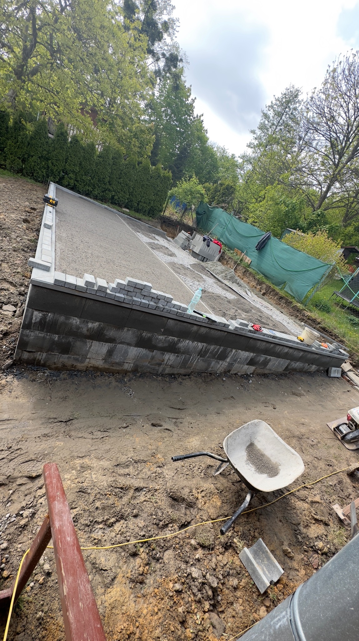 Budowa fundamentu pod taras z betonowych bloczków, widoczny piasek, poziomica laserowa i taczka z piaskiem na placu budowy otoczonym zielenią.