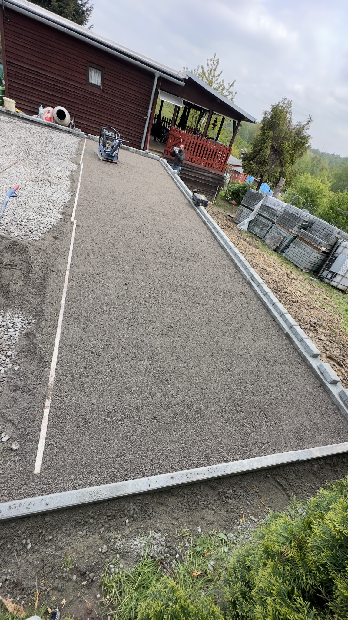 Przygotowanie podłoża pod ścieżkę z kostki brukowej: ubijanie zagęszczarką, widoczne obrzeża i stos kostki brukowej w tle.