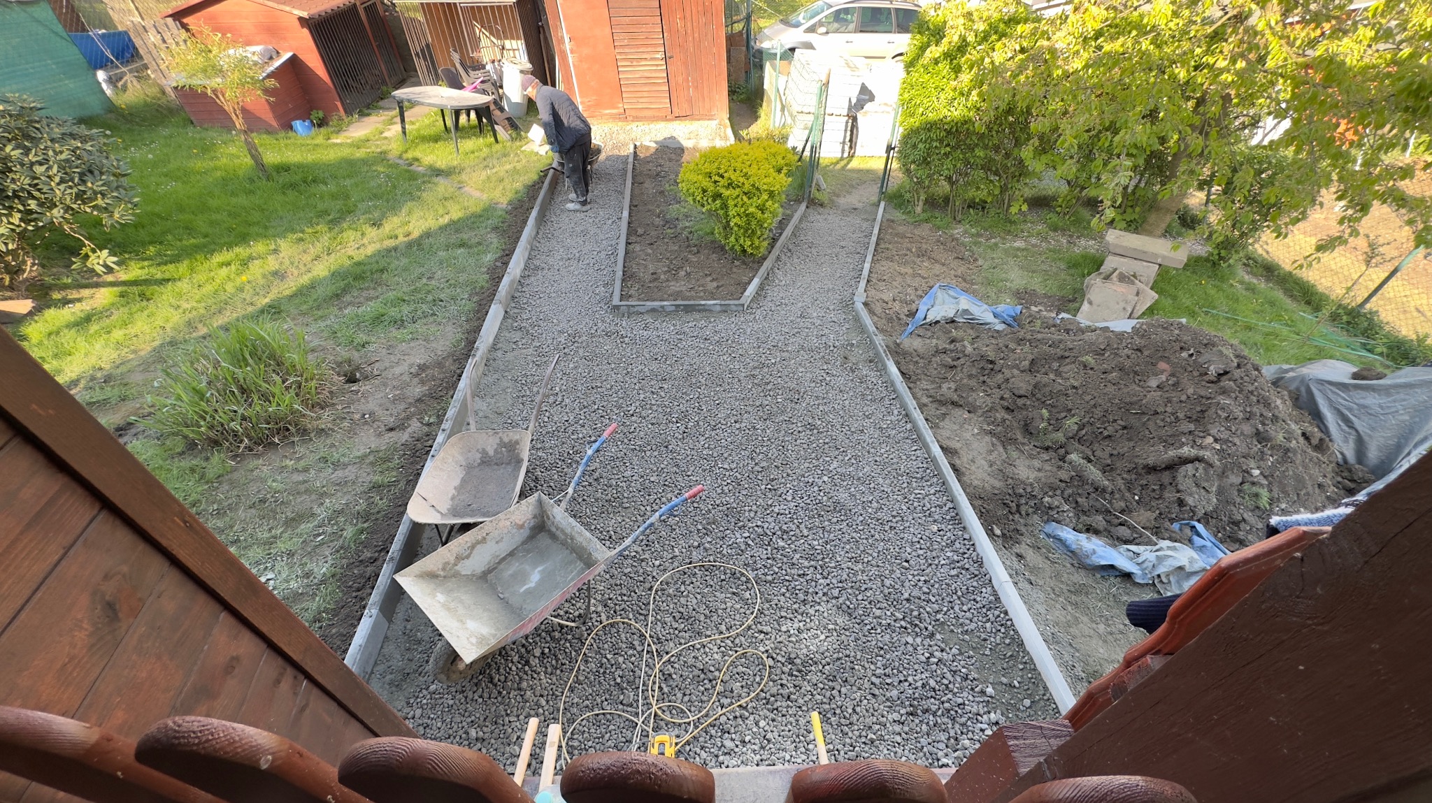 Budowa ścieżki w ogrodzie z wysypanym żwirem, widok z góry na dwie taczki, narzędzia i pracownika ubijającego kamienie