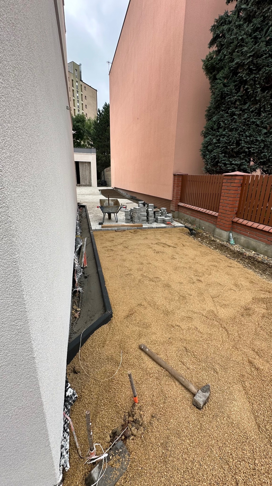 Przygotowanie terenu pod kostkę brukową: wyrównany piasek, obrzeża, paleta z kostką, taczka i młotek brukarski na budowie między budynkami.