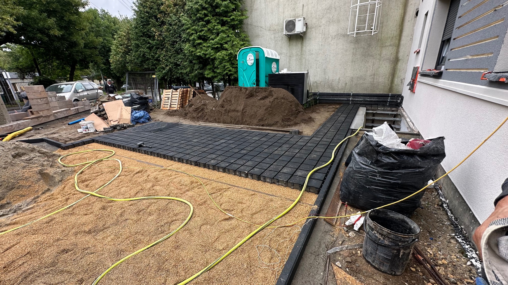 Układanie czarnej kostki brukowej na podsypce z piasku na zewnątrz budynku, widoczny przenośny WC i przygotowany teren pod dalsze prace.